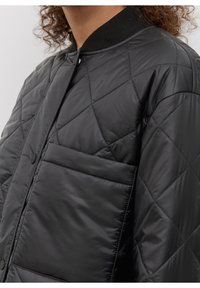 Sort quiltet bomberjakke med høj krave, frontlukning med trykknapper og en lomme med klap på venstre side. Glat, skinnende tekstur.