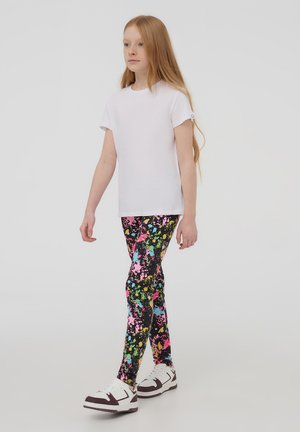 T-shirt bianca a maniche corte abbinata a leggings con motivo a macchie colorate nelle tonalità di nero, rosa, giallo e blu, e scarpe da ginnastica bianche.