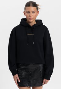 Zwarte cropped hoodie met een gouden logo aan de voorkant, voorzien van een trekkoordcapuchon en losse mouwen, gecombineerd met een zwarte leren rok met textuur.