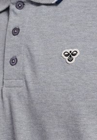 Polo gris en tissu texturé avec trois boutons foncés et un petit logo d'abeille brodé en crème et noir sur le côté gauche.