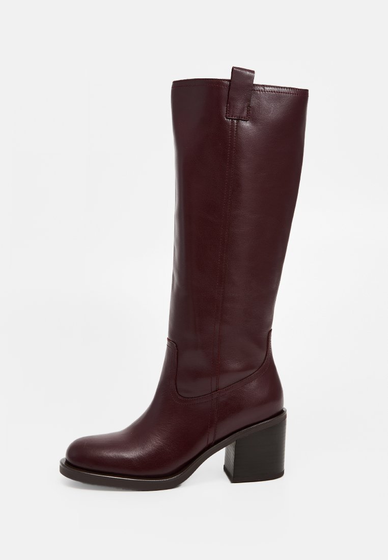 Bota de cuero burdeos hasta la rodilla con punta cuadrada, tacón ancho y un lazo trasero para ponérsela fácilmente. Textura suave y detalles de costura mínimos.