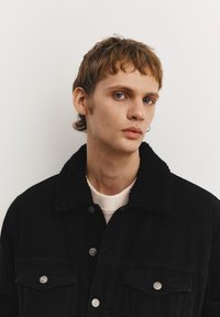 Mlajša oseba s kratkimi svetlo rjavi lasmi, modrimi očmi, oblečenimi v črno jakno s krznatim ovratnikom, rahlo obrnjena v stran na enostavni ozadju.