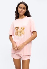 Femme aux longs cheveux noirs portant un short et un t-shirt roses assortis avec trois chiots marron clair et le texte "GUESS".