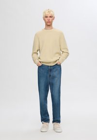 Beidzis teksturēts adīts džemperis, zilas džinsas bikses un gaišas krāsas sporta apavi. Modelis stāv taisni ar rokām kabatās pret neitrālu fonu.