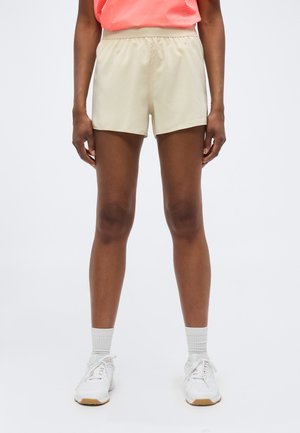 Personne portant un short de sport beige, des chaussettes blanches, des baskets blanches et un top sans manches corail sur un fond blanc uni.