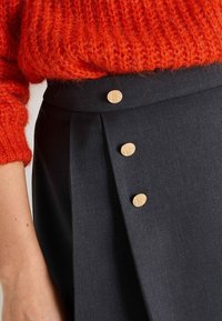 La falda negra presenta tres botones de madera en una cinturilla plisada, combinada con un suéter esponjoso de un brillante color naranja. La vista en primer plano resalta las texturas.