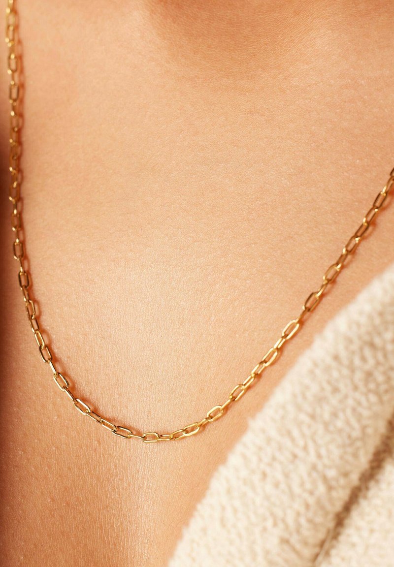 Collier en chaîne dorée avec des maillons allongés et entrelacés. La surface a une finition lisse, reposant sur une peau nue avec un arrière-plan texturé.