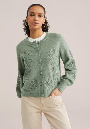 Femme portant un cardigan boutonné vert sauge avec des motifs floraux en relief et un pantalon beige, posant avec une main dans la poche.