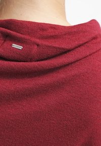 Gros plan sur le cou d'une personne portant un pull en maille rouge doux à col roulé avec une petite étiquette métallique de la marque cousue sur le col.