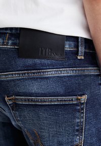 Jeans en denim bleu foncé avec coutures apparentes et un patch en cuir noir portant l'inscription "LTB", portés avec un haut blanc.