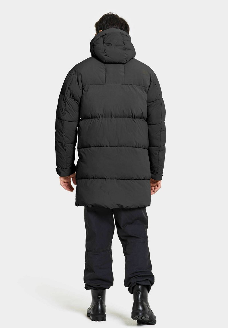 Schwarze Pufferjacke mit Kapuze, mit gestepptem Design, verdeckten Reißverschlüssen und verlängertem Saum, kombiniert mit schwarzen Hosen und robusten Stiefeln.