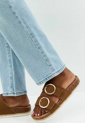 Personne portant un jean bleu clair et des sandales marron avec deux grandes boucles dorées, montrant les pieds et les jambes inférieures.