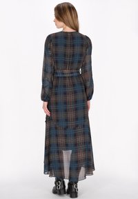 Lange, doorzichtige, geruite jurk in donkerblauw, met een ceintuur in de taille, lange mouwen en een gelaagde, vloeiende onderrand. Zwarte enkellaarzen met gespen.
