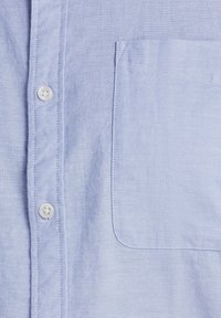 Himmelblaues Hemd mit Knöpfen, das eine Brusttasche und einen strukturierten Stoff aufweist. Es hat weiße Knöpfe und sichtbare Nähdetails.