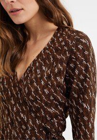 Brun mönstrad omslagsblus med V-ringning, som har vita och orange prickar, texturerad tyg och en knytning i midjan.