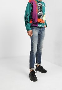 Färgglad tröja med grafiska mönster i teal, lila och orange. Bärs med ljusblå jeans och svarta sneakers, med uppvikta ärmar.