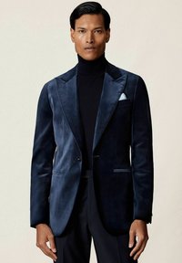 Námornícky sametový blejzer so saténovými klopami, s jedným gombíkom, bočnými vreckami a bielym vreckovým štvrtkom, nosený cez čierny rolák.