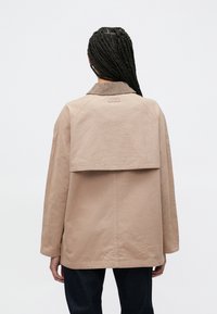 Smėlio spalvos per didelis švarkas su ribuotu apykakliu, siūlių detalėmis ir sluoksniuotu nugaros dizainu. Medžiaga atrodo tekstūruota ir tvirta.