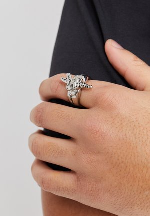 Silver ring with a sword design, featuring a textured band and engraved text. Ring sits on a hand resting against a dark fabric.