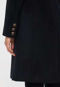 Must must coat, millekudumine, sile tekstuur ja neli dekoratiivset kuldset nuppu mansetil lisadetailina.