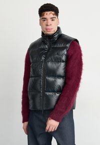 Svart pufferväst med hög krage och dragkedja fram, med glänsande yta och horisontell quiltning. Bärs över en bourgognefärgad fluffig tröja.