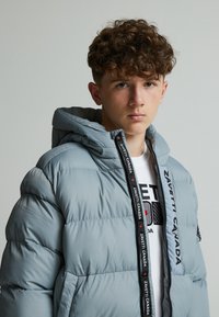 Veste rembourrée gris clair avec capuche, dotée d'une fermeture éclair et d'un logo noir le long de la fermeture. Texture lisse et design matelassé.