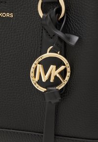 Czarna skórzana torba z teksturowaną powierzchnią, z złotym okrągłym zawieszką z logo "MK" na środku oraz detalem z czarnej skóry na pasku.