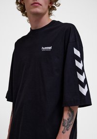 Zwarte katoenen T-shirt met korte mouwen, met een wit logo op de borst en witte chevronpatronen op de mouwen.