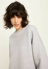 Junge Person mit welligem, dunklem Haar, die einen lockeren, hellgrauen Sweatshirt trägt, frontal schaut und mit einem leichten Lächeln vor einem einfarbigen Hintergrund steht.