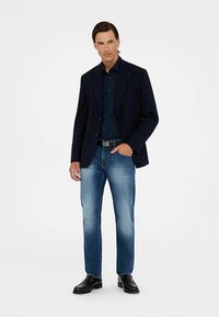 Blazer azul marino sobre una camisa oscura con botones, combinado con jeans azul claro y zapatos negros. Presenta un logo sutil en el blazer.