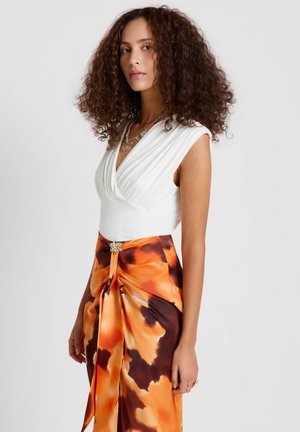 Donna con capelli ricci che indossa una canotta bianca e una gonna a motivi arancioni e marroni con una spilla decorativa in vita.