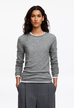 Femme aux longs cheveux bouclés foncés portant un pull gris par-dessus une chemise blanche et un pantalon gris, debout les mains dans les poches.
