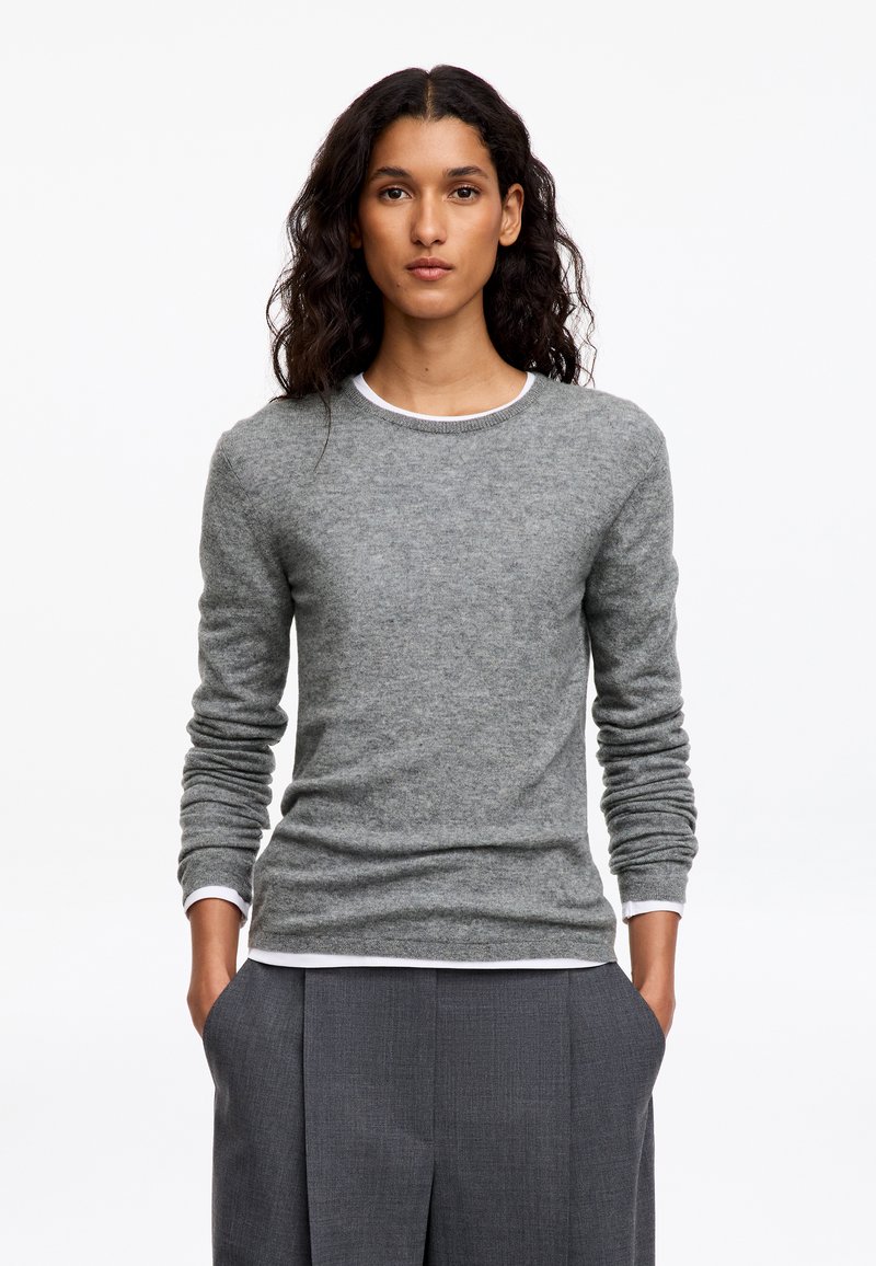 Femme aux longs cheveux bouclés foncés portant un pull gris par-dessus une chemise blanche et un pantalon gris, debout les mains dans les poches.