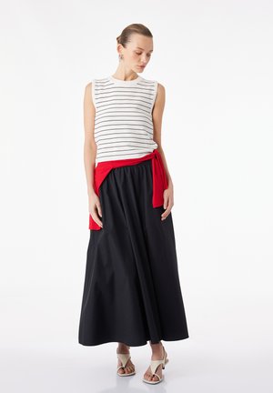 Vrouw draagt een mouwloze witte top met zwarte strepen, lange zwarte rok, rode trui geknoopt in de taille en witte open teen hakken, staand tegen een witte achtergrond.
