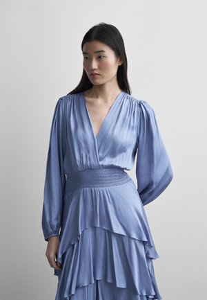 Mujer de cabello largo y oscuro que lleva un vestido azul claro de manga larga con escote en V profundo y falda de capas, mirando hacia un lado sobre un fondo liso.