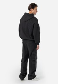 Sort oversized hoodie med et struktureret logo på ryggen, parret med løstsiddende sorte cargo-bukser og hvide sneakers.