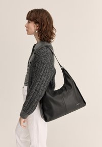 Žena oblečená v tmavosivom pletenom kardigane a bielych nohaviciach nesie veľkú čiernu koženú kabelku na rameno s oblým horným okrajom.