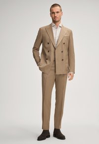 Giacca marrone doppiopetto con rever a punta, abbinata a pantaloni coordinati e una camicia chiara, completata da stivali alla caviglia in suede marrone scuro.