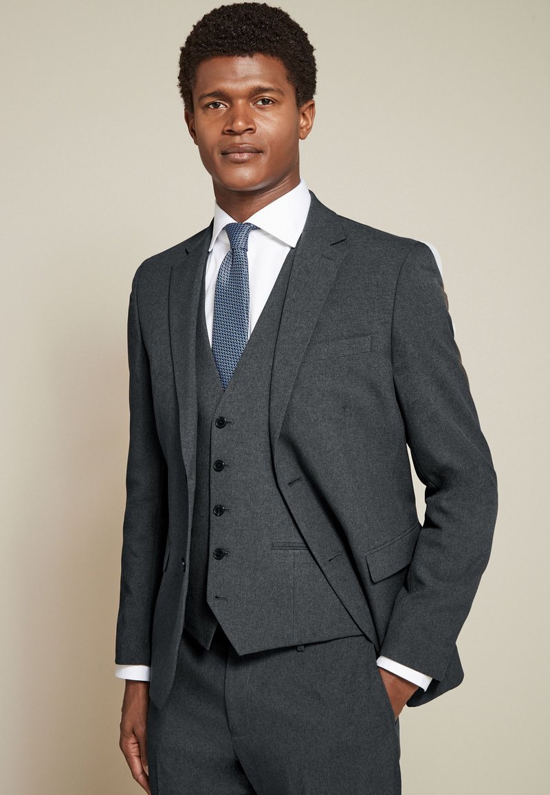 Homme portant un costume trois pièces gris foncé avec une chemise blanche et une cravate bleue à motifs, debout avec une main dans la poche.