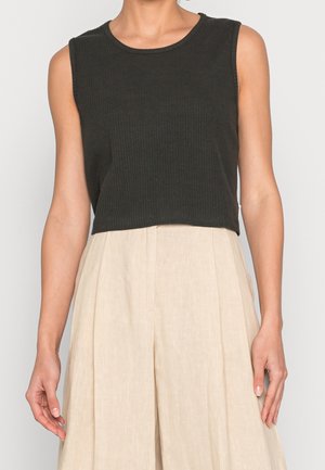 Mouwloze zwarte geribbelde cropped top gecombineerd met beige geplooide, wijde hoge taille broek bij een staand persoon.