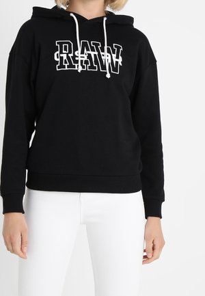 Zwarte hoodie met witte trekkoorden en de tekst "RAW" op de borst, gedragen door een persoon met witte broek, staand tegen een witte achtergrond.