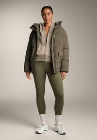 Oliva zelena puhovka, bež kapucar z zadrgo, oprijete olivno zelene legice, bele superge z detajli in svetle nogavice.