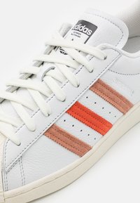 Sneaker bianca in pelle con tre strisce orizzontali in suede arancione e marrone, dotata di suola testurizzata e design con lacci.