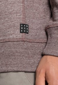 Sudadera en tejido marrón jaspeado, con puños y dobladillo acanalados, y una etiqueta negra con la inscripción "DNM WRKRS" cosida en un lado.