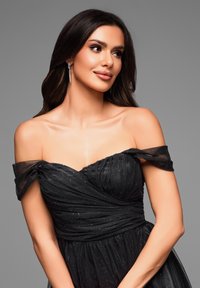 Robe de soirée noire avec des manches en tulle décolletées, un corsage froncé et une jupe fluide ornée de paillettes discrètes. Accentuée par des boucles d'oreilles scintillantes.