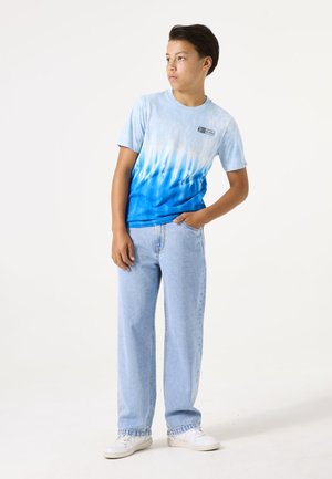 Camiseta de manga corta azul con efecto tie-dye, de degradado claro a oscuro; combinada con unos vaqueros de corte holgado y lavado claro, y zapatillas blancas.