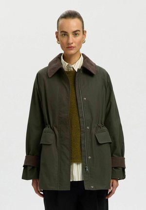 Donna che indossa una giacca verde scuro con colletto e polsini in velluto a coste marrone, sovrapposta a una camicia a righe e un maglione oliva, in piedi davanti a uno sfondo grigio.