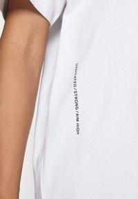 Bílý bavlněný tričko s vertikálním černým textem "DEDICATED/STRONG/AIM HIGH" na boku. Hladká textura, uvolněný střih.