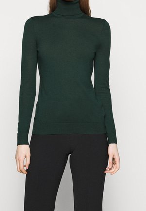 Kobieta w ciemnozielonym dopasowanym golfie i czarnych spodniach, ukazana od szyi do bioder na jednolitym tle.