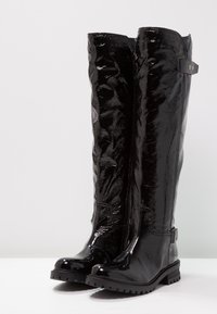 Czarne kozaki za kolano wykonane z błyszczącej skóry lakierowanej, z fakturowaną powierzchnią, płaską gumową podeszwą oraz metalowymi ozdobami przy górnej krawędzi i obcasie.